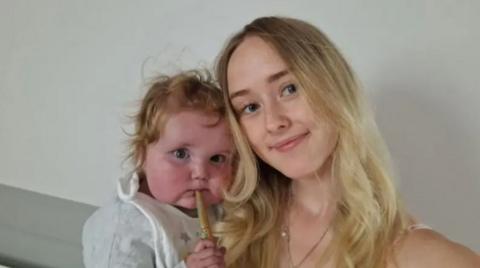 A woman with blond hair alongside a baby with blonde curly hair and a stick in his mouth, wearing a white top and bib