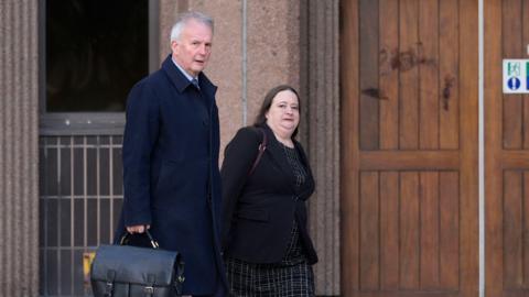 Esther Baker walking into court, she wears a black patterned dress under a black blazer. She has long brown hair. She walks alongside a man who has short grey hair. He wears a long blue coat and carries a black bag.