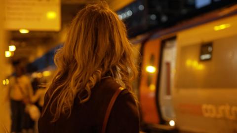 The back of a woman with long blonde hair and a black cardigan sat on a station platform at night with a train in the background.