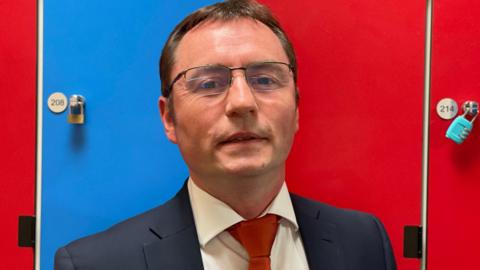 Dominic Burke pictured wearing glasses, a suit and tie looking at the camera and standing in front of red and blue lockers