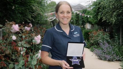 A woman in dark blue nurse scrubs with white piping on the sleeves and collar. She has light brown hair tied back and is standing in a garden with flowers and bushes around her. She is holding a cushioned box with a medal in it, shaped like a blue star, that reads "Cavell".