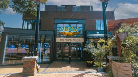The entrance to The Alban Arena, with large glass windows and doors. Trees, plants, and two tall street lamps are near the entrance.
