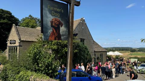 An historic stone pub building under blue skies with rolling fields in the distance. A pub sign with a picture of a fox red labrador on it reads 'The Farmer's Dog'. There is a queue of people outside the pub door. 