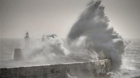 A huge wave crashes over a breakwater and lighthouse, with grey skies overhead and stormy seas in the background