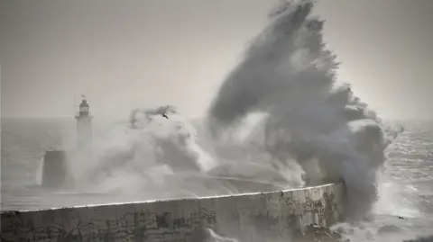 A huge wave crashes over a breakwater and lighthouse, with grey skies overhead and stormy seas in the background