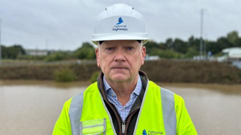 A man wearing a national highways white hard hat and high vis vest standing in front of a brown river