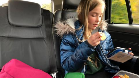 A child sits in the back on a car looking at a hand held electronic device and eating a snack from their lunch box. She is wearing a school uniform and winter coat
