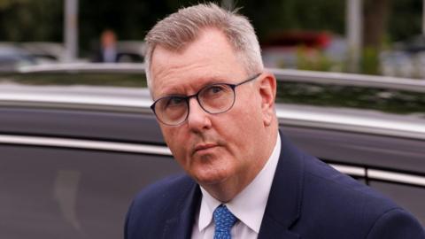 A man with short, mostly grey hair, is exiting a car. He is wearing black-rimmed glasses, a blue suit jacket, white shirt and blue-patterned tie. The car behind him is black. Out of focus, in the background, are other vehicles, lampposts and shrubbery. 