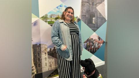 Molly Watt - with long brown hair, tied back, with a dark blue and white dress and a denim jacket standing outside the BBC Radio Berkshire studios with Bella, a black labrador 