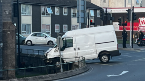 A picture of a large white van which has crashed into a roundabout. It has damaged the railings and police are pictured in the background.