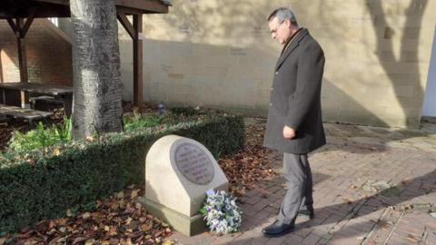 A man, Councillor Mark Ieronimo, wearing a smart grey coat and trousers bows his head towards a memorial with a wreath on. The granite memorial is beneath a tree and autumn leaves surround it. 