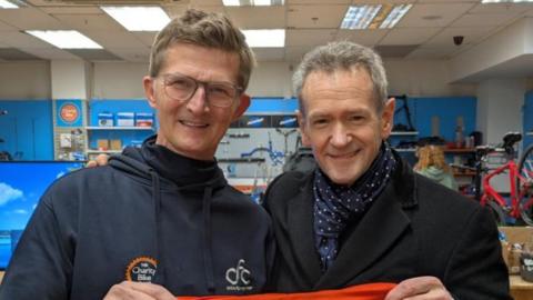Alexander Armstrong is on the right and has grey hair with a black coat and blue and white scarf. He is holding one side of a red top that reads "one big thank you" in white writing. On the left is Michael who has brown hair and glasses and is wearing a blue navy hoodie, and is holding the other side of the top.