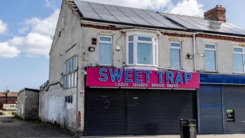 Sweet Trap in Shotton Colliery. The unit is based on the ground floor of an end-of-terrace building and the shutters are pulled down. The sign is bright pink with blue lettering and advertises cakes, shakes, book and vapes. The upper level has a central bay window with two smaller windows on either sides, with white vertical blinds. The cream paint is coming off the building in parts. There are solar panels on the roof.
