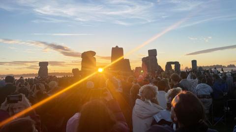 The sun is framed by two stones of Stonehenge which then reveal a strong ray of light. There are many people gathered watching the sunrise.