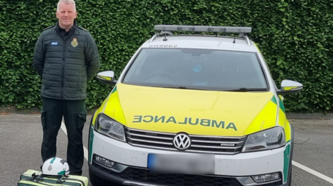 Gavin Palin wearing a dark coloured NWAS uniform and coat stood by an ambulance car with kit