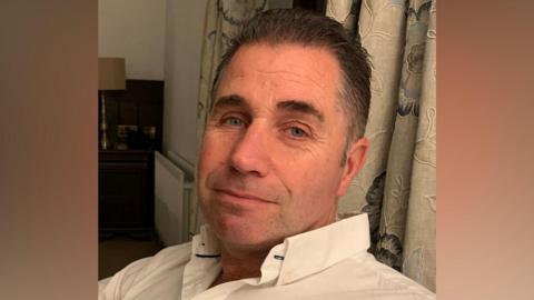 A middle-aged man with dark short hair that us greying at the sides. He is looking toward the camera and is wearing a white button down shirt. He is sitting in front of some cream curtains with a floral design. 