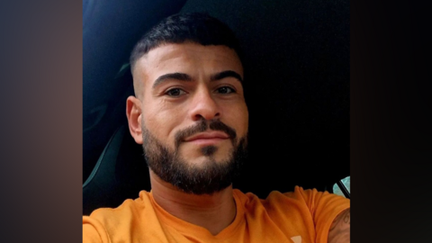 Andrew Robson, sitting in a car, smiling. He has short, black hair and a beard and is pictured wearing an orange top.