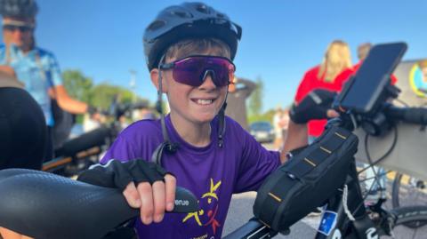 A smiling Dexter is preparing to set off on his cycle fundraiser to Paris. He is wearing a purple T-shirt for one of the charities he is supporting, Falconi Hope, a purple pair of sports glasses and a helmet. He is about to get on his bike. Other people can be seen in the background.