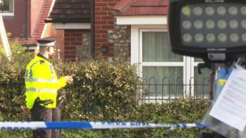 A police officer in a reflective yellow coat and uniform, stood behind blue and white police tape with a red brick house behind him. 