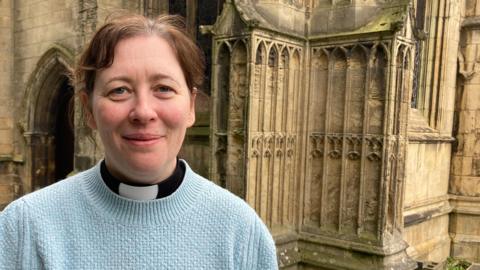 The Rev Becky Lumley wearing a light blue jumper over a vicar's black shirt and white dog-collar stands in front of a corner of a medieval stone church. She has brown hair, tied back, and is smiling.