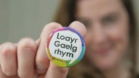 Ruth Keggin-Gell, who has brown hair, holding up a badge that says Loayr Gaelg rhym.