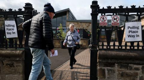 Members of the public attend a polling station in the village of Quarter to cast their votes. A man is walking through a gate towards the polling station and a woman is walking out of the gate.