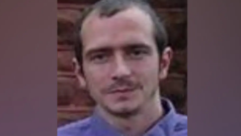 A photograph of Paul Cunliffe. He is standing in front of a brick wall and wears a shirt. He has short brown hair and a thin moustache.
