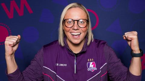 Blond-haired Vic Burgess smiling and cheering wearing glasses and her purple Loughborough Lightning training top. 