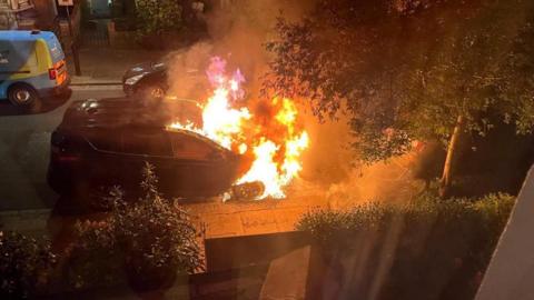 The front half of a car parked on a residential street is seen engulfed in flames. The picture appears to have been taken from inside a neighbours house at night time.