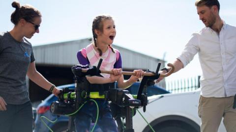 Brook stands, holding onto a horizontal wooden bar. The exoskeleton is black and runs down her legs, with a green bar around her middle. Brooke, whose dark hair is tied in pigtail plaits, smiles, and has a man standing on one side of her and a woman on the other.