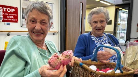 Two older women with short grey hair are sitting down. One is wearing a light green cardigan and top, and is holding a knitted pink heart while smiling. The other woman is wearing a blue and white top with a blue cardigan and is holding a cream coloured knitted heart. There is a brown wicker basket with a blue handle between them, filled with more knitted hearts