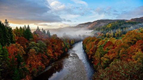 An autumnal scene from Killiecrankie in Perthshire. A river runs through the middle of the scene. On either side, trees coloured orange, yellow, red and green are under a blue sky. There is cloud above the trees on the left side with the light from the sunrise just poking through. In the distance, a layer of fog covers part of the trees and river in front of several hills