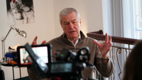 Dave Singleton, wearing a beige top with short cropped grey hair, waves his arms as he is interviewed in front of a camera