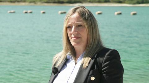 Susan Davy pictured in front of a large reservoir on a sunny day. She is wearing a light blue blouse with a dark coloured blazer. She has straight long dark blonde hair. 