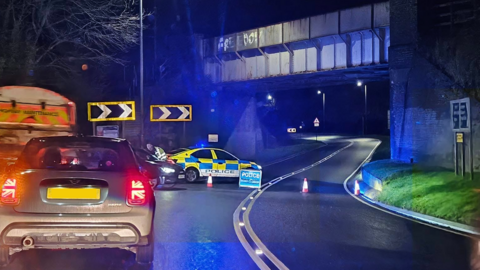 The scene of the crash with a police sign and cones to block the road, as well as a police vehicle.