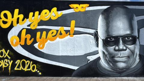 Mural of Carl Cox on baordwalk by stadium. Cox is a black man with shaved head. he has been painted wearing sunglasses in black and white with yellow letters either side saying Oh yes oh yes, and The Prodigy