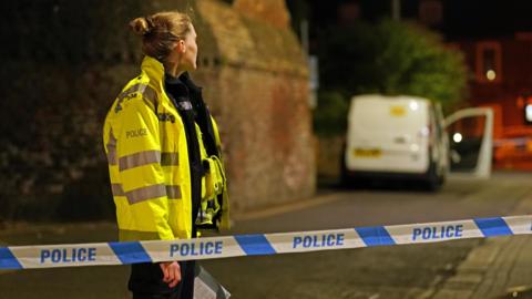A female police officer standing next to police tape in an area which has been cordoned off. It is dark and there is a white van with the door open in the background.