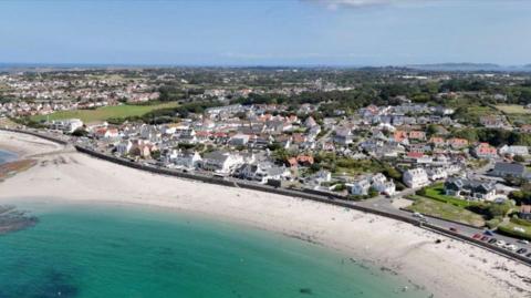 A drone image of Guernsey with the sea on the bottom with sand and houses further back. 