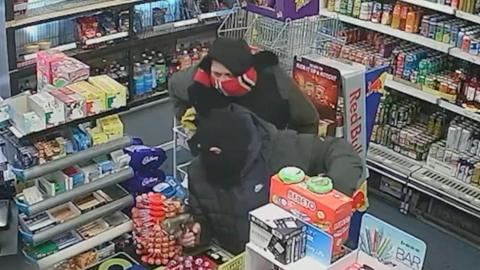A still from CCTV footage showing two men standing at the counter of a convenience shop. One is wearing a black jacket and a black balaclava, while pointing an imitation hand gun. The other is wearing a black hooded jacket with a red scarf covering his face.