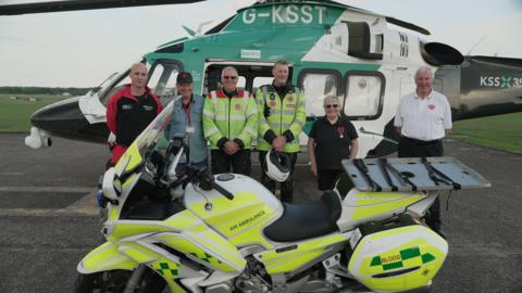 Five men and a woman are stood infront of a helicopter belonging to Air Ambulance Charity Kent Surrey Sussex. Five of them are volunteers, one of them is a Doctor for the air ambulance. Infront of them is an air ambulance motorbike. 