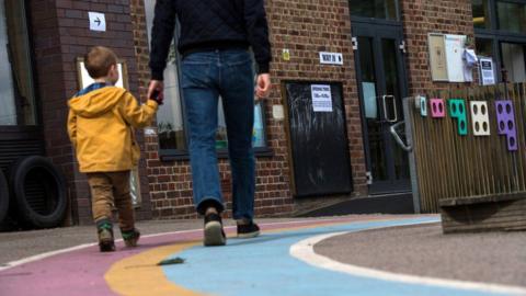 A small boy wearing a yellow raincoat holds his dad's hand as they walk into a polling station in a primary school.
