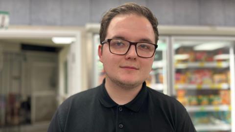 Kieran Essex looks directly at the camera, unsmiling. He has a black polo shirt and black glasses. He is standing in a convenience store with a fridge full of food. 