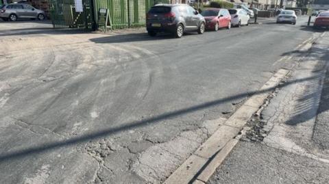 View of Seavy Road showing cracks in the tarmac on the path and road. There are  cars parked along one side of the road, as well as an entrance gate to premises.