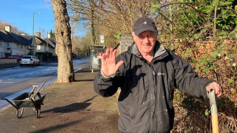 A man in a waterproof coat and cap on the side of a road holding a spade and waving, with a wheelbarrow and broom nearby