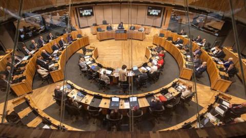 Senedd members seated at their desks in the parliament's circular chamber.