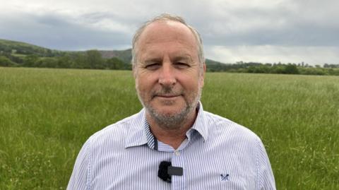 Mike Rigby is standing in a green field wearing a blue and white pin-striped shirt. He has short grey hair and a short beard.