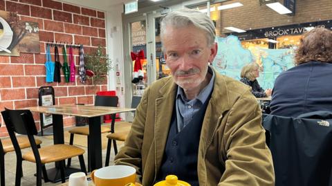 Graham sits at a table with a pot of tea at The Canterbury Bakery wearing a waistcoat suit and corduroy jacket