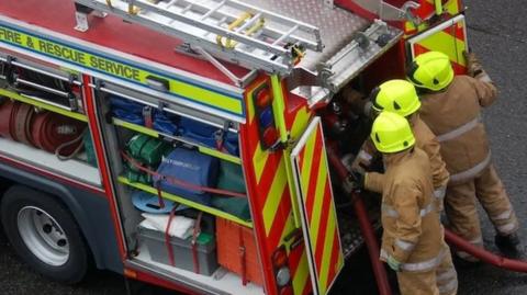 Three firefighters wearing yellow helmets and brown uniforms have their backs to the camera as they handle hoses attached to the back of a red fire engine. Compartments are open showing various pieces of equipment. A ladder is on the roof.