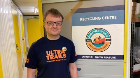 A man is standing facing the camera. He is wearing a dark navy or black short-sleeved T-shirt and rectangular eyeglasses with dark frames. The T-shirt has prominent orange text. Visible text on the shirt includes “ULTRA TRAILS” in large orange letters. Smaller text beneath reads “YORKSHIRE WOLDS ULTRA”. Behind the person is a large vertical structure resembling a container. It has a logo bearing the "PRELOVED SPORTS" name.