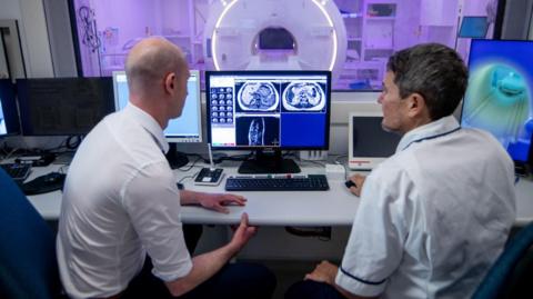 Two men sit in front of the MRI Simulator machine, which enables cutting-edge imaging technology for radiotherapy treatment, at Leeds Teaching Hospitals NHS Trust.